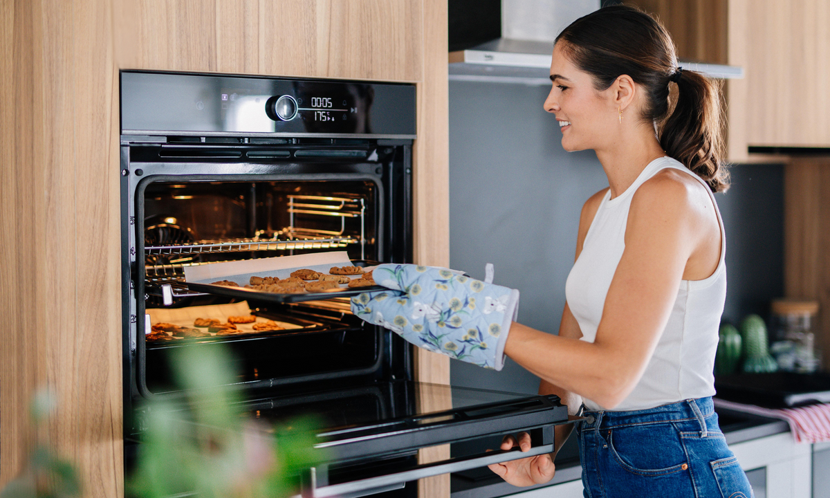 A woman with a white singlet is cooking using her Beko Oven in her kitchen, she is putting cookies in the oven