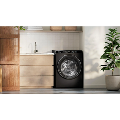 Front view of the Haier 15/9kg washer dryer combo in a black finish, in a modern laundry room with wooden cabinetry and a tree in a pot plant nearby.