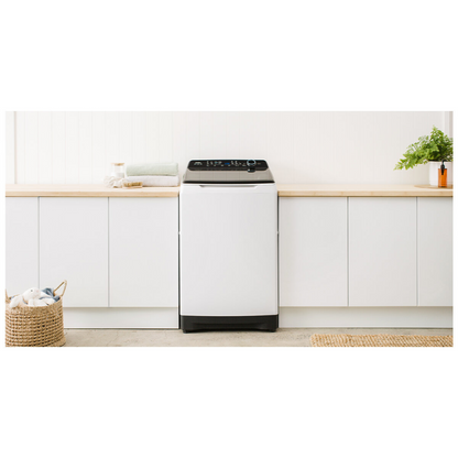 Front view of the Haier 12kg top load washing machine in a white finish with the lid closed, in a modern laundry set up with white cabinetry and wooden benchtops, with folded towels and a pot plant on the wooden benchtops.