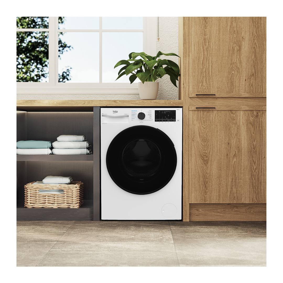 A Beko washer & dryer combo in white with a black door, integrated into a modern laundry setup with wooden cabinetry and open shelving. Folded towels and a wicker basket are neatly arranged nearby, and a potted plant sits on the bench above, creating a clean and stylish laundry space with natural light streaming in from the window.