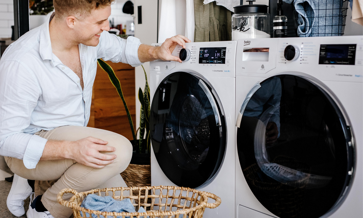 A man bends down to adjust the settings on his white Beko Washing machine and Dryer in his laundry