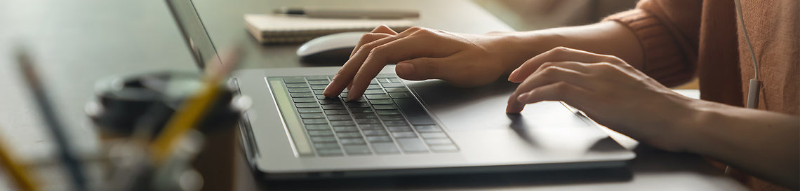 A person in a light brown sweater is typing on a laptop keyboard, with a notebook and pen nearby, illuminated by natural light.