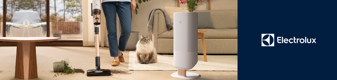 Someone using an Electrolux petpro plus vacuum cleaner in a modern living room setting, a pot plant spilt over with soil on the floor. A cat on the carpet and a wooden table nearby. 