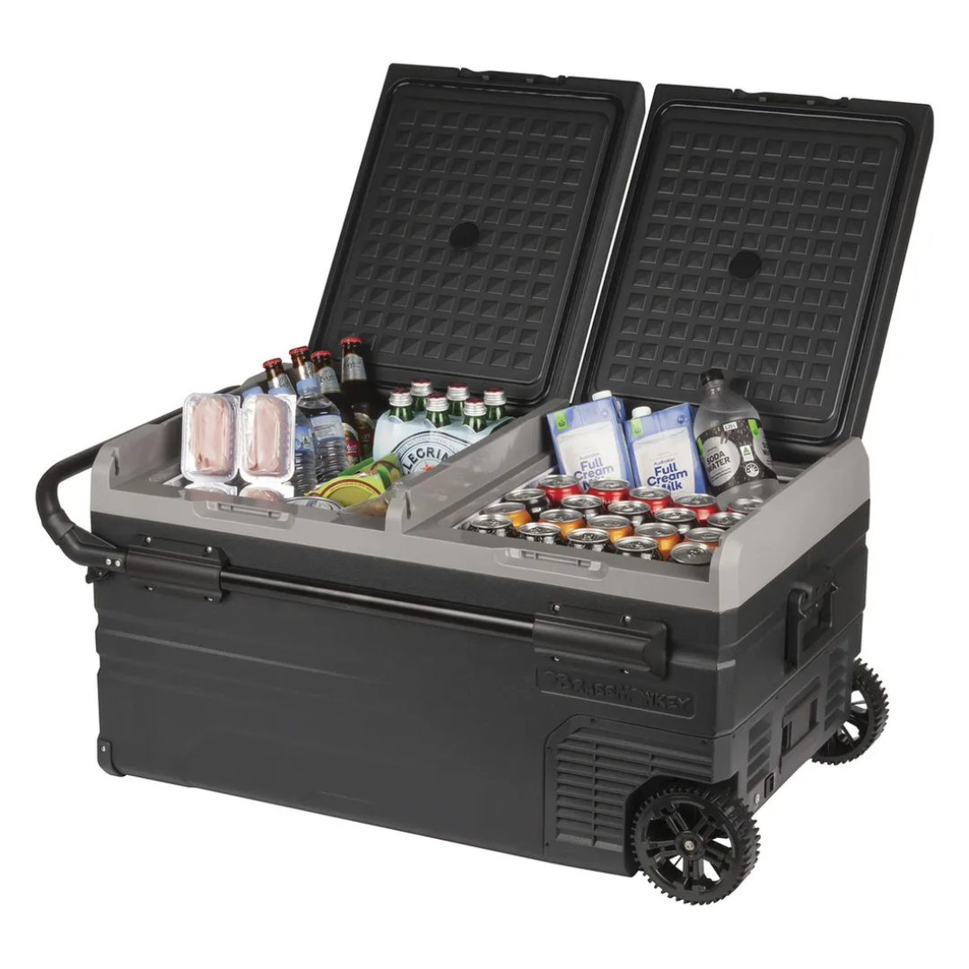 Angled view of the Brass Monkey dual lid portable fridge/freezer with wheels and telescopic handle, stocked with drinks and food, ideal for camping, road trips, or outdoor adventures.  