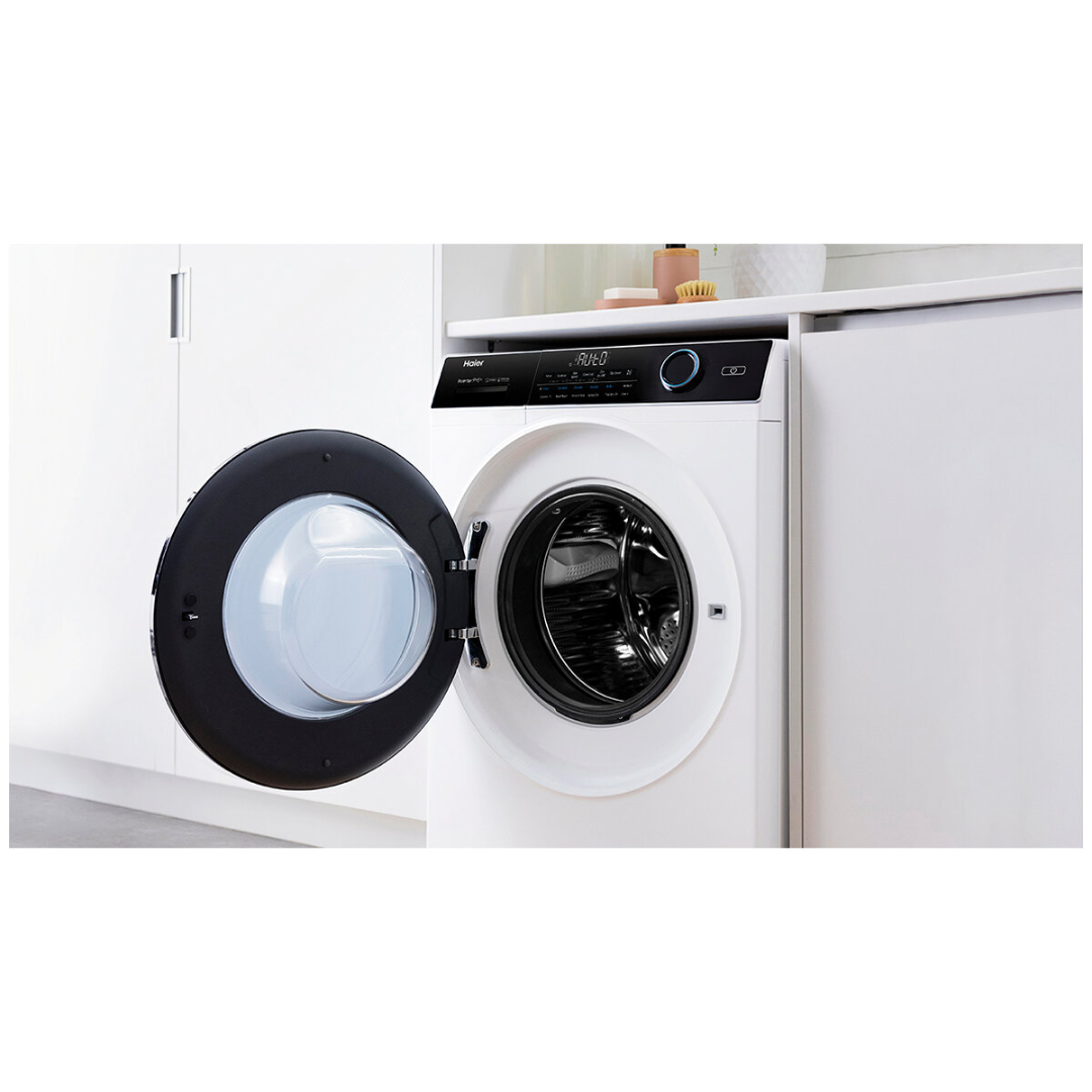 Angle view of the Haier 9/5kg washer dryer combo in a white finish with the door open, in a modern laundry room with white cabinetry. 