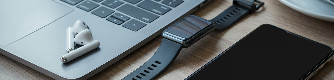 A close-up view of a modern workspace with a grey laptop, wireless earbuds, a smartwatch, and a smartphone neatly arranged on a wooden surface.