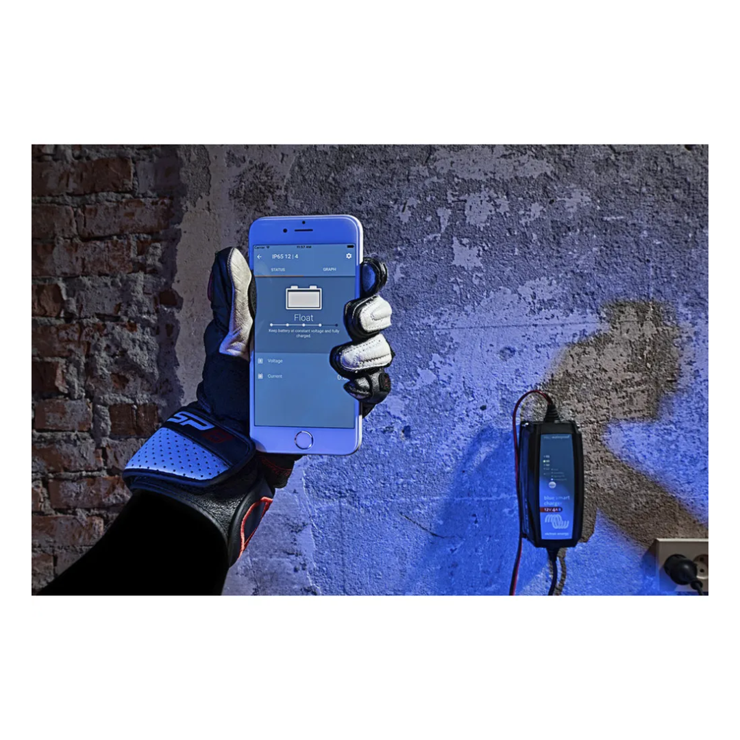 Gloved hand holding smartphone showing Bluetooth app for Victorian Professional Blue Smart charger, mounted on wall and actively monitoring battery charging status. 