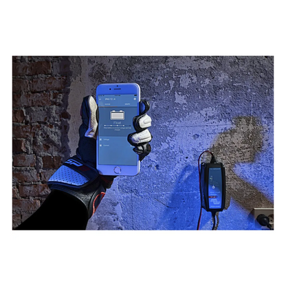 Gloved hand holding smartphone showing Bluetooth app for Victorian Professional Blue Smart charger, mounted on wall and actively monitoring battery charging status. 