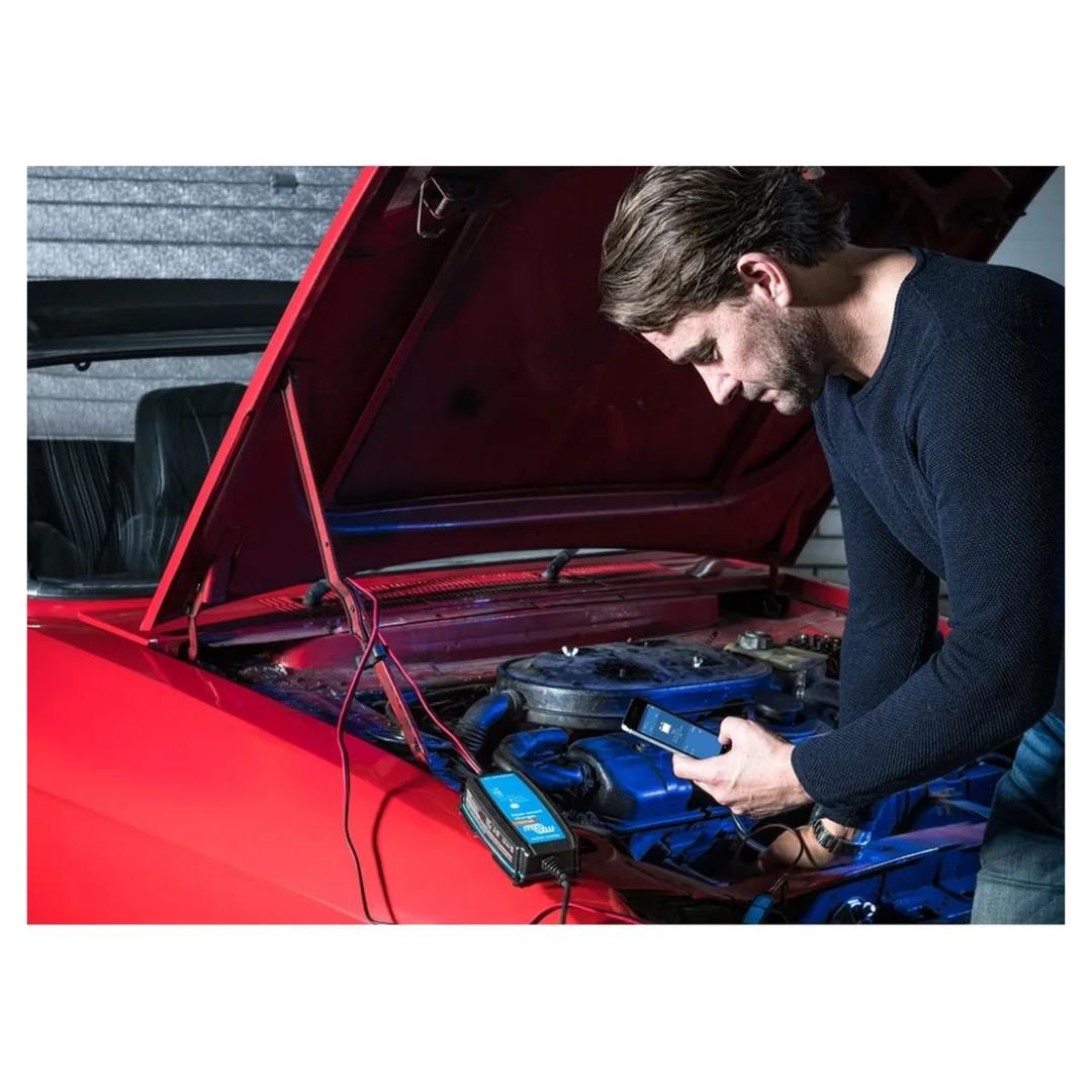 Man using smartphone to monitor car battery with Victorian Professional Blue Smart Charger connected under bonnet, showcasing Bluetooth control and smart charging features. 