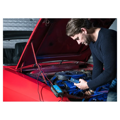 Man using smartphone to monitor car battery with Victorian Professional Blue Smart Charger connected under bonnet, showcasing Bluetooth control and smart charging features. 