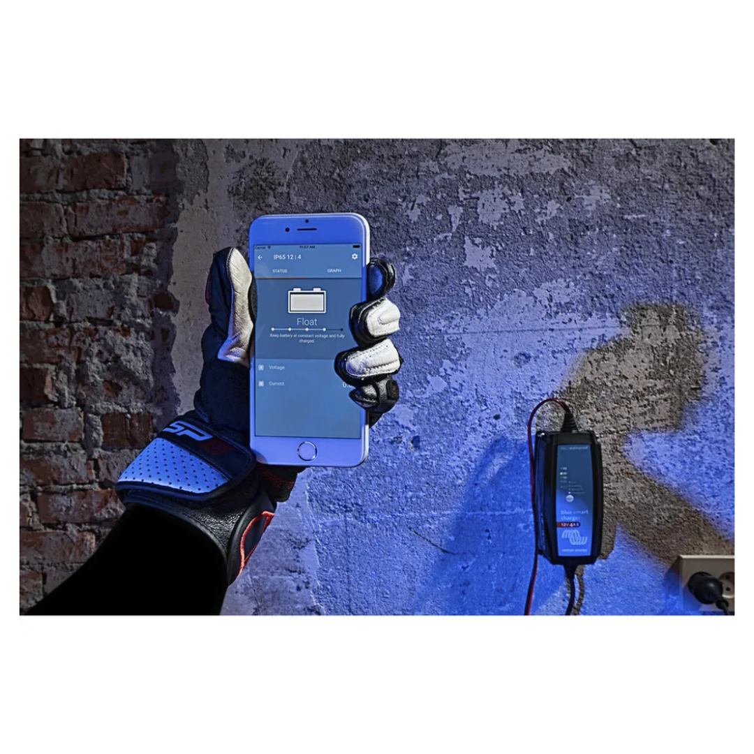 Gloved hand holding smartphone showing Bluetooth app for Victorian Professional Blue Smart Charger, mounted on wall and actively monitoring battery charging status.