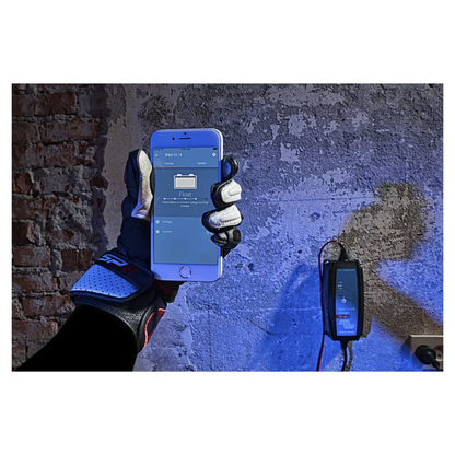 Gloved hand holding smartphone showing Bluetooth app for Victorian Professional Blue Smart Charger, mounted on wall and actively monitoring battery charging status.