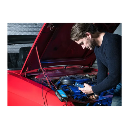 Man using smartphone to monitor car battery with Victorian Professional Blue Smart Charger connected under bonnet, showcasing Bluetooth control and smart charging features. 