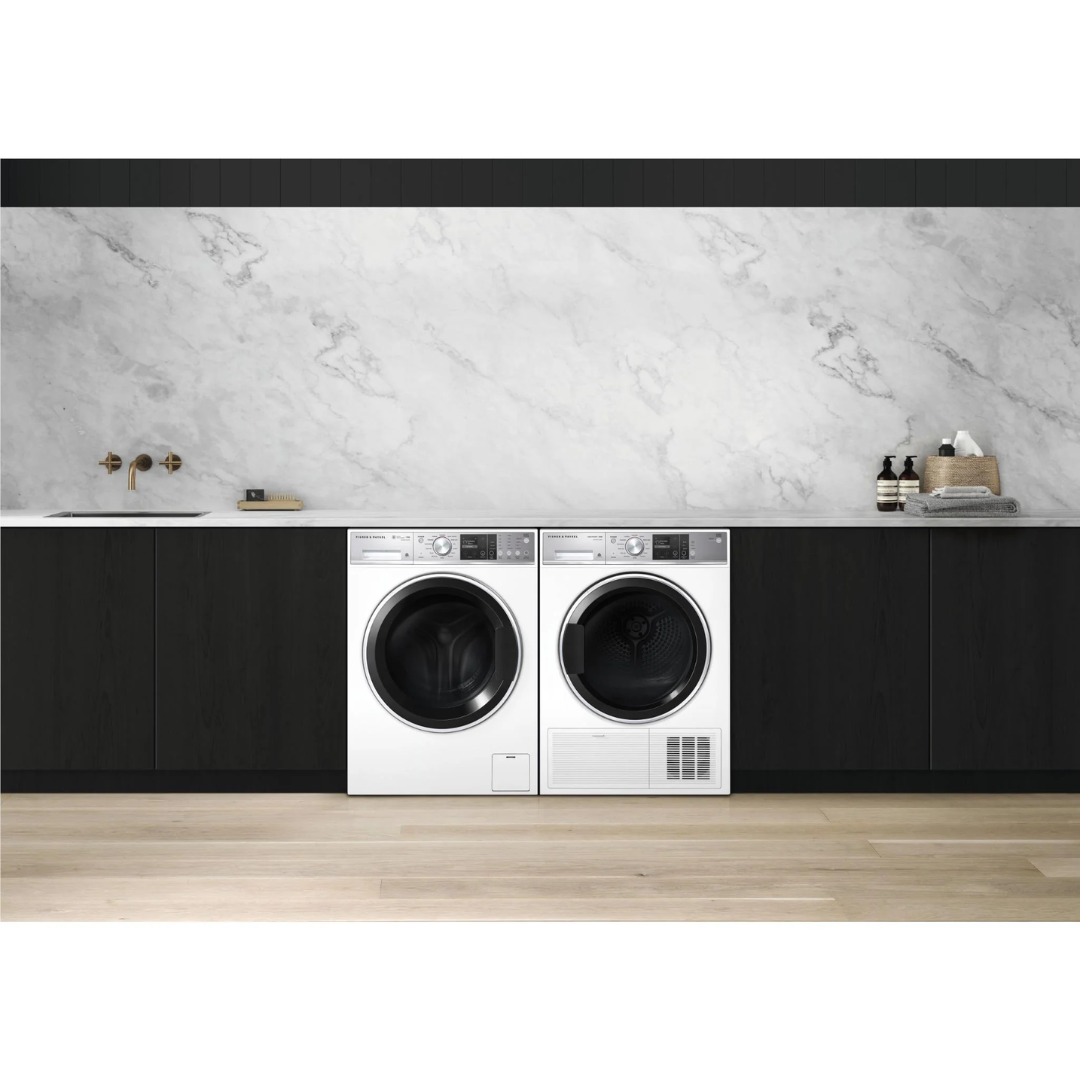 Front view of the Fisher & Paykel 11kg front load washing machine in white, alongside a white Fisher & Paykel dryer, in a modern laundry setting, with black cabinetry besides them, a marble benchtop, detergents nearby and a tap & sink on the left side. 