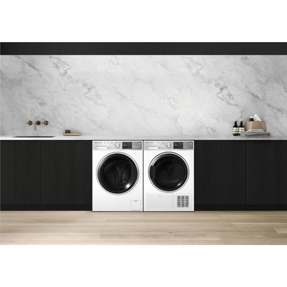 Front view of the Fisher & Paykel 11kg front load washing machine in white, alongside a white Fisher & Paykel dryer, in a modern laundry setting, with black cabinetry besides them, a marble benchtop, detergents nearby and a tap & sink on the left side. 