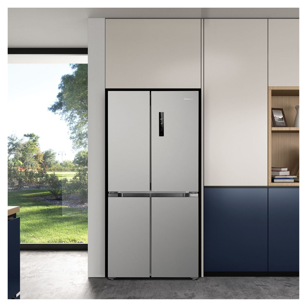Front view of a 496L French Door Fridge with all doors closed in a modern kitchen setting with wooden cabinetry and open shelving. A picture frame and books are neatly arranged nearby. Natural light streaming though the window. 